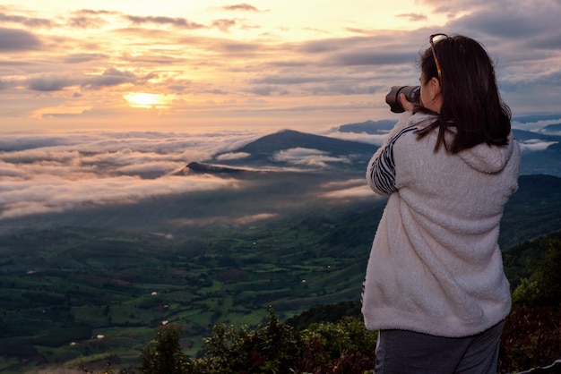 Frauentouristen verwenden eine DSLR-Kamera, die im Winter bei Sonnenaufgang auf einem hohen Aussichtspunkt im Nationalpark Phu Ruea, Provinz Loei, Thailand, die Naturlandschaft des Sonnennebelbergs fotografiert