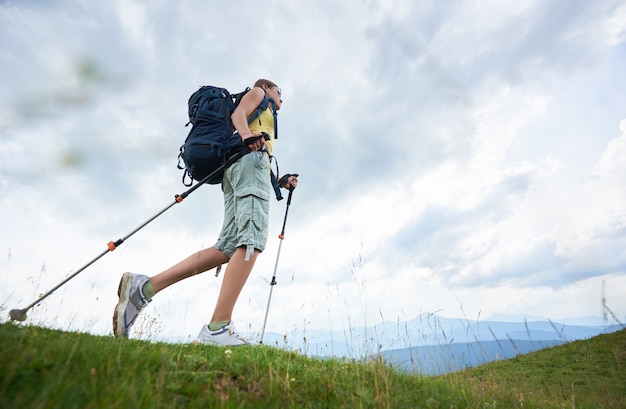 Frauenwanderer, der auf grasbewachsenem Hügel wandert, Rucksack tragend, mit Trekkingstöcken in den Bergen