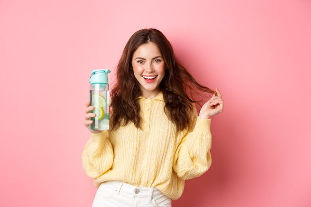 Freche attraktive Frau, die zuversichtlich in die Kamera schaut, hält persönliche Flasche mit gesundem Zitronenwassergetränk und steht gegen rosa Wand.