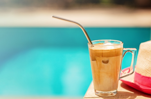 Fredo de café helado contra el agua azul clara de la piscina con sombrero de paja