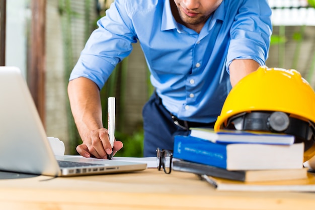 Foto freelance - arquitecto trabajando en casa en un diseño o borrador, en su escritorio hay libros, una computadora portátil y un casco o casco