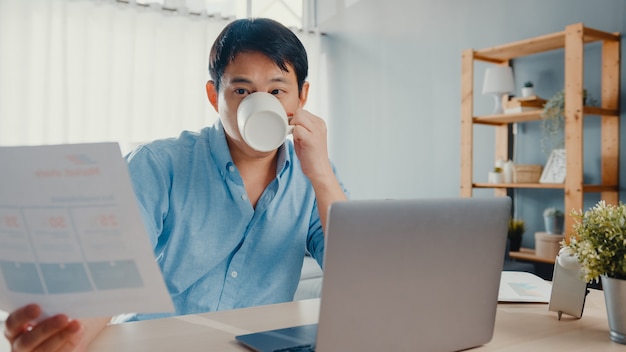 Freelance Asia guy casual wear usando laptop y tomando café en la sala de estar de la casa.