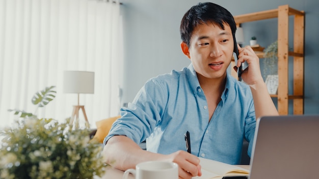 Freelance Asia Guy Freizeitkleidung mit Laptop-Talk auf Handy im Wohnzimmer im Haus.