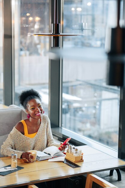 Freelancer alegre. Freelancer alegre usando seu smartphone e bebendo café enquanto está sentado em um restaurante