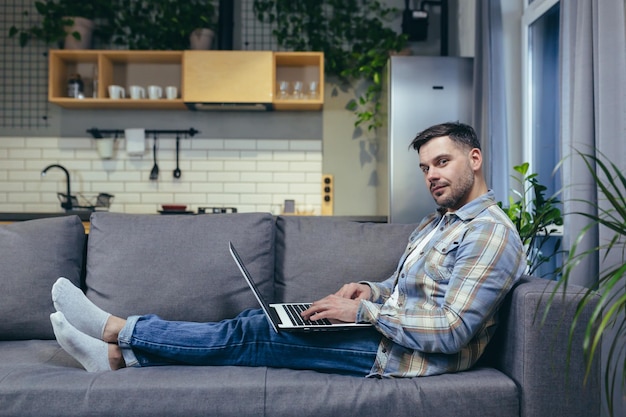 Freelancer Apuesto joven trabajando en la computadora portátil en casa Acostado en el sofá con ropa de casa