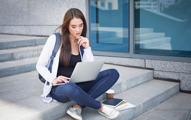 Freelancer asiático pensativo al aire libre. Joven morena en las escaleras de un área urbana, bebiendo café y trabajando con una laptop en línea. Concepto de estilo de vida y tecnología.