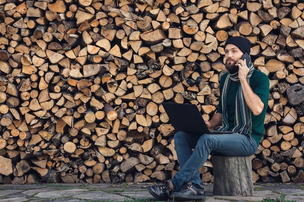 Freelancer barbudo está trabalhando no laptop e falando ao telefone contra o fundo da parede de lenha