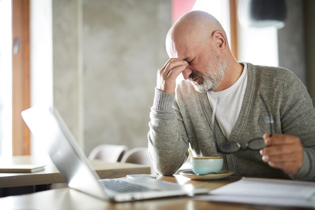 Freelancer calvo sênior cansado com barba esfregando os olhos enquanto tira os óculos no café