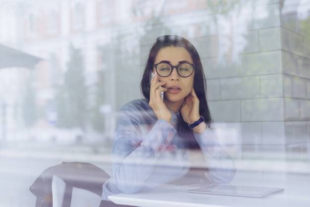 Freelancer de chica joven ocupada en camisa azul y gafas está negociando por teléfono