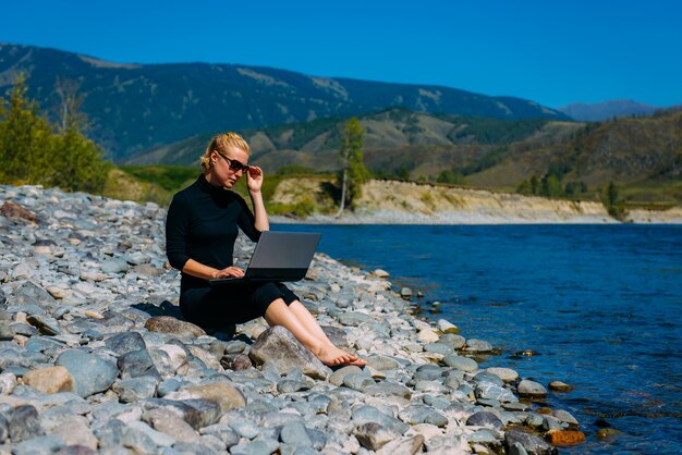 Freelancer com laptop sobre a natureza. Blogger hipster viajante negócios senhora menina bonita trabalha ao ar livre. Blondy jovem de preto sentado ao lado do rio da montanha.