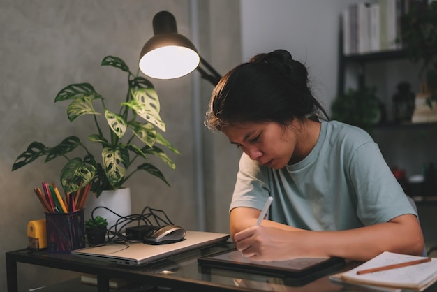 Freelancer de designer gráfico de mulher asiática trabalhando horas extras à noite usando tablet para desenhar trabalho de arte do conceito de casa