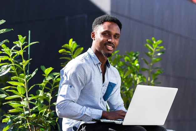 Freelancer de homem negro trabalhando online usando laptop sentado no banco do lado de fora do edifício moderno do escritório no parque urbano da cidade na rua. um estudante afro-americano feliz em um ambiente casual ao ar livre digitando no teclado