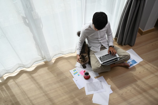 Freelancer de jovem trabalhando no laptop com seu gato enquanto estão sentados juntos em um quarto moderno.