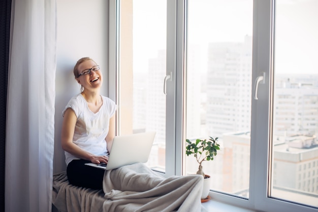 Freelancer de mulher adulta jovem feliz de óculos, sentado no fundo da janela. Loira rindo trabalhando no laptop. Local de trabalho no parapeito da janela. Conceito de escritório em casa.