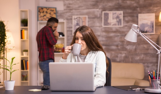 Freelancer de mulher branca, trabalhando no laptop na sala de estar e desfrutando de uma xícara de café. Namorado falando ao telefone e comendo batatinhas no fundo.