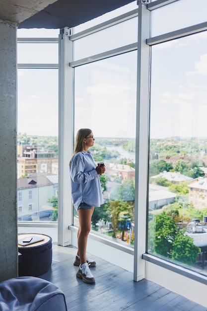 Freelancer de mulher moderna falando ao telefone enquanto está em uma grande janela e bebendo café O conceito de uma mulher moderna e bem-sucedida Jovem em um escritório aberto