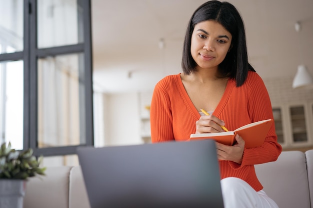 Freelancer de mulher pensativa usando laptop tomando notas assistindo cursos de treinamento trabalhando em casa