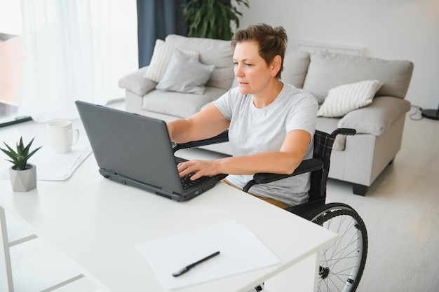 Freelancer em cadeira de rodas usando laptop perto de notebook e papéis na mesa