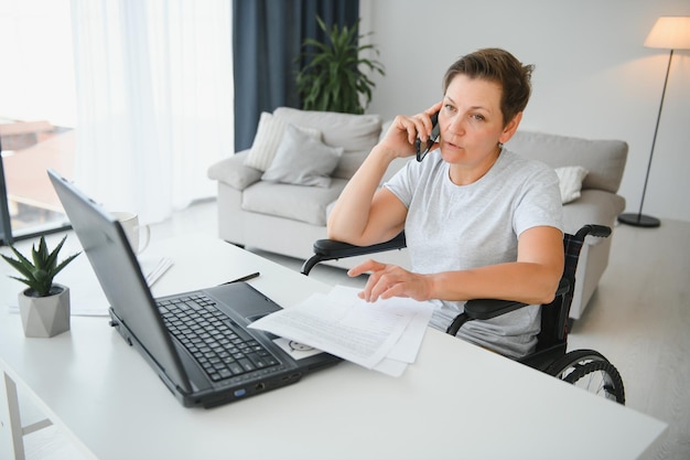 Freelancer em cadeira de rodas usando laptop perto de notebook e papéis na mesa