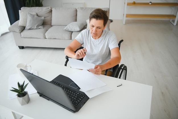 Freelancer em cadeira de rodas usando laptop perto de notebook e papéis na mesa