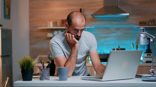 Freelancer estresado por la sobrecarga de trabajo quedándose dormido mirando en la computadora portátil sentado en la cocina de casa. Empleado remoto agotado durmiendo una siesta en la silla que se despierta trabajando en la PC usando una red de tecnología moderna