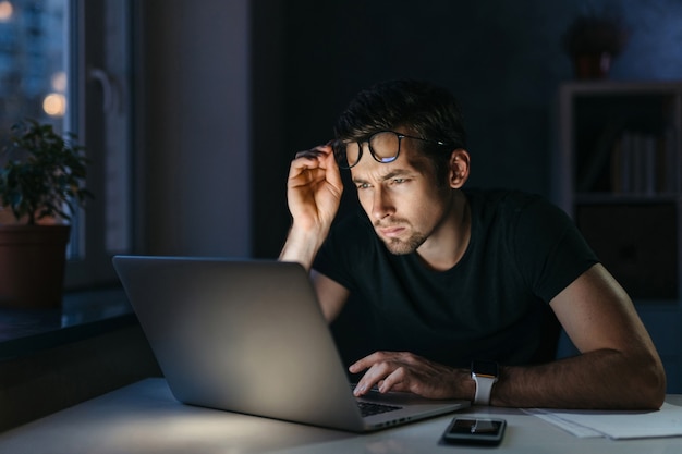 Freelancer estudiante hombre cansado con exceso de trabajo trabaja de forma remota con una computadora portátil por la noche, sosteniendo gafas siente fatiga ocular