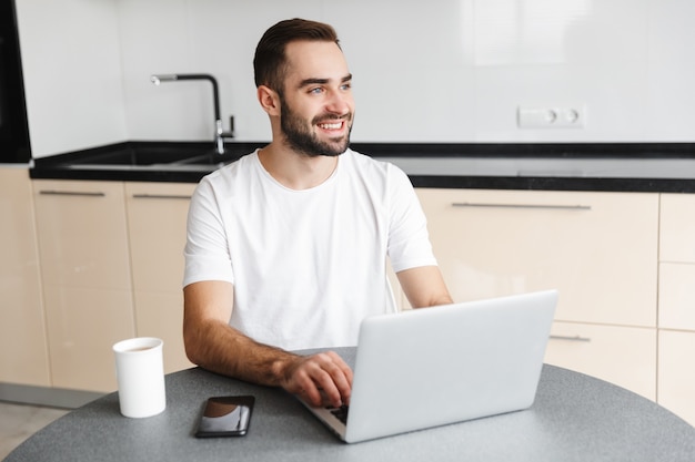 Freelancer feliz homem bonito sentado à mesa da cozinha, trabalhando em um computador laptop