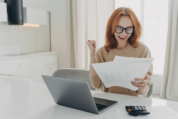 Freelancer femenino comprueba los cálculos aprieta el puño mira felizmente los documentos en papel