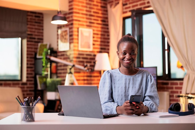 Freelancer feminino bem sucedido trabalhando com celular no escritório em casa moderno. Gerente de SMM afro-americano sorridente usando smartphone e laptop, mensagens e olhando para a câmera