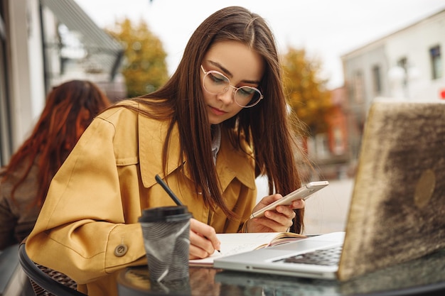 freelancer feminino fazendo anotações no caderno e usando o celular enquanto trabalhava remotamente no café