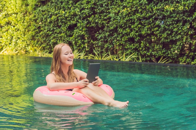 Freelancer feminino jovem sentado perto da piscina com seu laptop no hotel navegando em seu smartphone. Ocupado nos feriados. Conceito de trabalho distante. Copie o espaço para o seu texto