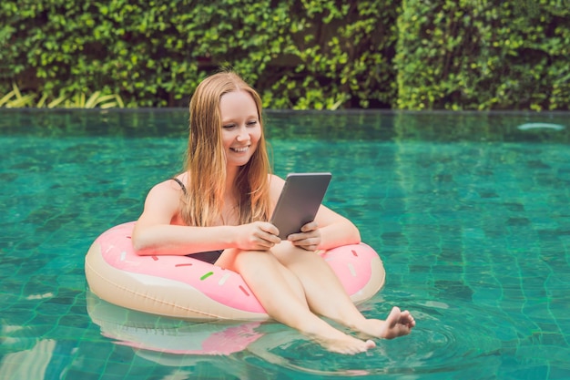 Freelancer feminino jovem sentado perto da piscina com seu laptop no hotel navegando em seu smartphone. Ocupado nos feriados. Conceito de trabalho distante. Copie o espaço para o seu texto.