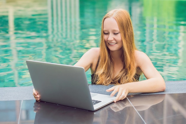Freelancer feminino jovem sentado perto da piscina com seu laptop. Ocupado nos feriados. Conceito de trabalho distante. Copie o espaço para o seu texto