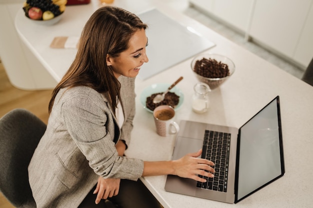 Freelancer feminino sorridente usando seu laptop e tomando café da manhã em casa.