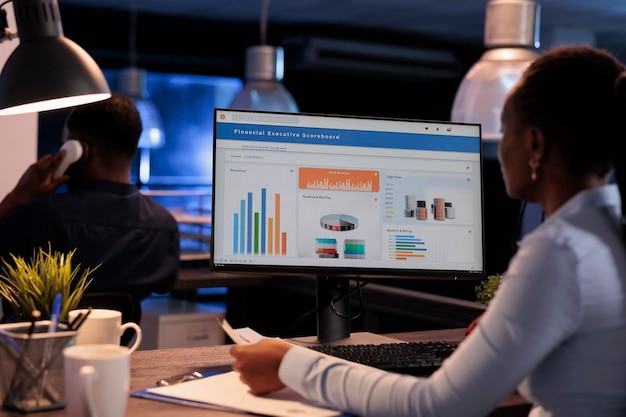 Freelancer feminino trabalhando com gráficos de estatísticas no monitor do computador, analisando dados da empresa na papelada. Usando informações on-line para criar relatórios de pesquisa antes do prazo, à noite após o expediente.