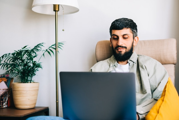 Freelancer hombre caucásico trabajando en casa, sentado en un sillón y usando la computadora portátil.