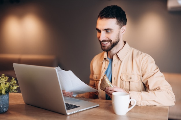 Freelancer hombre comiendo sándwich, tomando café y reuniéndose con el jefe por videollamada