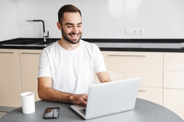 Freelancer hombre guapo feliz sentado en la mesa de la cocina, trabajando en la computadora portátil