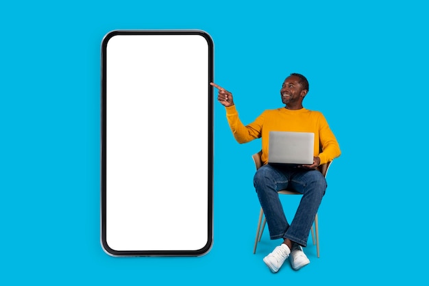 Freelancer de hombre negro feliz usando computadora apuntando a maqueta de teléfono inteligente