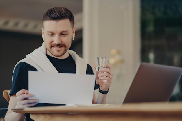 Freelancer homem olhando um documento na frente do laptop, sentado no escritório em casa em sua mesa de trabalho, segurando um copo de água e usando fones de ouvido, jovem empresário em roupa casual trabalhando remotamente de casa