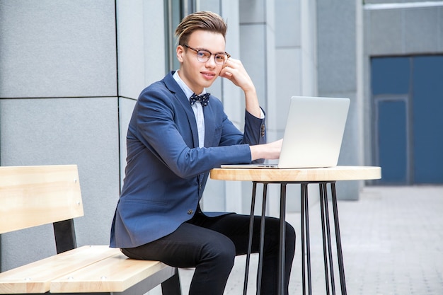 Freelancer jovem atraente com terno azul casual com laptop e café sentado e trabalhando. olhando para a câmera e sorrindo.