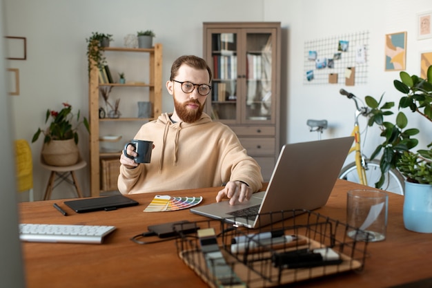 Freelancer jovem barbudo olhando para a tela do laptop enquanto toma um café e navega na rede em busca de informações sobre as novas tendências