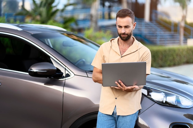 Freelancer latino usando laptop trabajando en línea parado cerca del auto. Trabajador haciendo diagnóstico de coche