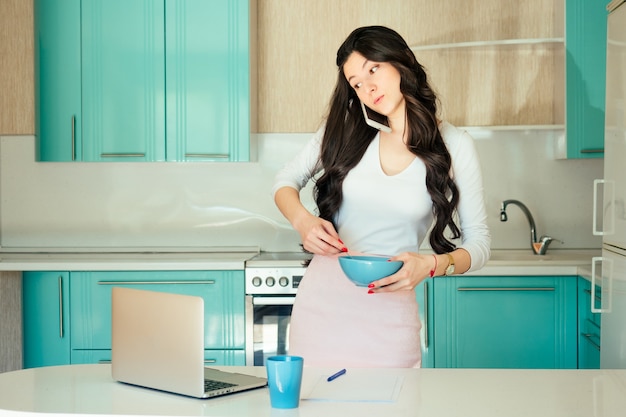 Freelancer linda jovem aluna em um vestido branco e cabelo preto trabalha em casa um laptop ee papéis Café da manhã na cozinha turquesa.