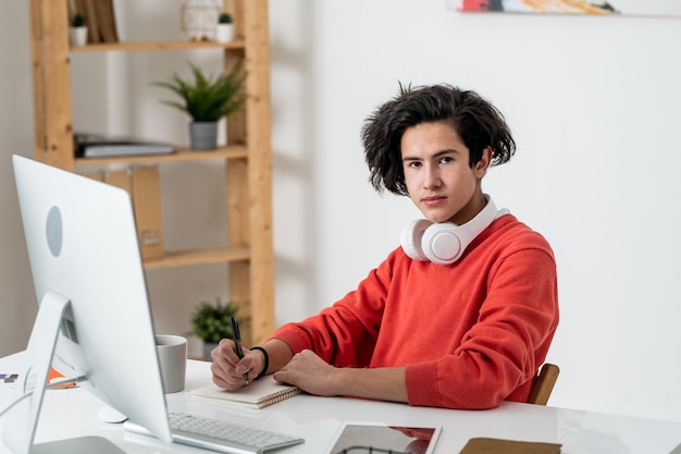 Freelancer masculino casual joven que le mira mientras toma notas en el escritorio frente a la pantalla de la computadora en casa