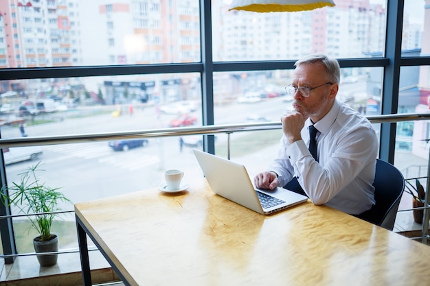 Freelancer masculino está trabajando en un café en un nuevo proyecto empresarial. Se sienta en una gran ventana en la mesa. Mira la pantalla de una computadora portátil con una taza de café.