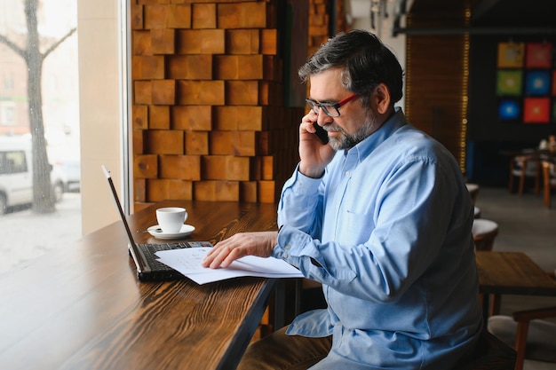 Freelancer masculino está trabalhando em um café em um novo projeto de negócios Senta-se em uma grande janela na mesa Olha para uma tela de laptop com uma xícara de café