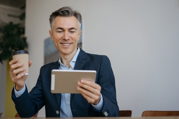 Freelancer de mediana edad guapo con tableta digital sosteniendo una taza de café trabajando desde casa