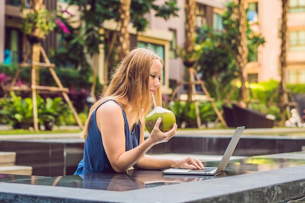 Freelancer mujer joven sentada cerca de la piscina con su computadora portátil en el hotel navegando en ella