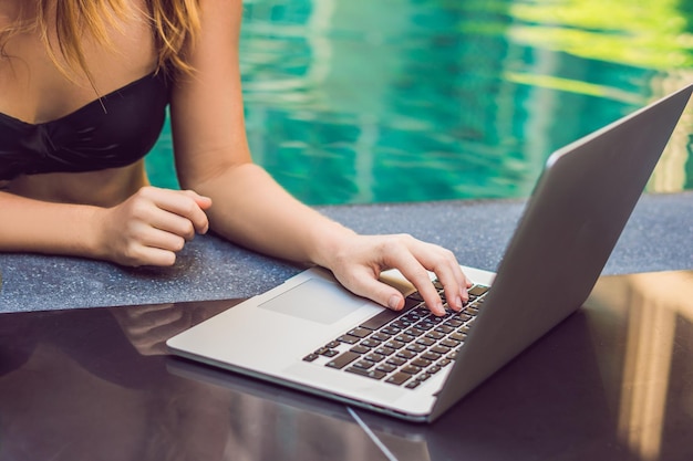 Freelancer mujer joven sentada cerca de la piscina con su computadora portátil en el hotel navegando en su teléfono inteligente. Ocupado en vacaciones. Concepto de trabajo distante. Copie el espacio para su texto.